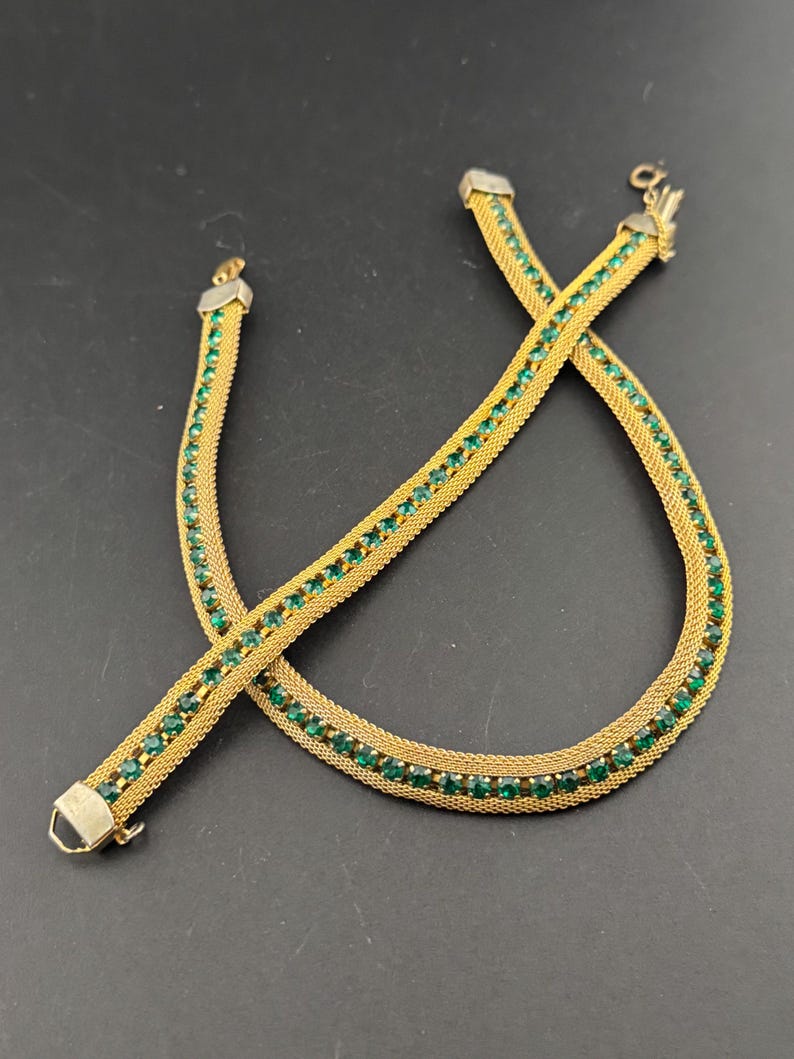 May include: A gold-toned necklace with a mesh design, adorned with rows of small, emerald-green gemstones. The necklace is laid out on a dark surface, showcasing its intricate details and clasp. The jewelry piece appears to be vintage or antique.