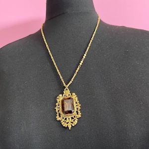 May include: A gold-toned necklace with a large, rectangular, amber-colored pendant. The pendant is set within an ornate, filigree frame. The necklace is displayed on a black mannequin against a pink background. The chain is delicate.