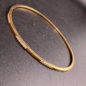 Peut inclure: Un bracelet jonc de couleur or avec un design rectangulaire. De petites pierres transparentes sont serties le long des sections rectangulaires du bracelet. Le bracelet est circulaire et semble être en métal.