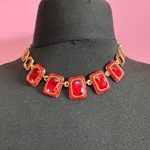 May include: A gold-toned necklace with large, rectangular red gemstones. The gemstones are set in a gold frame and are connected by small gold links. The necklace is displayed on a black mannequin head against a pink background.