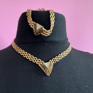 May include: Gold-coloured necklace and bracelet set. The necklace and bracelet feature a chain link design with a decorative centre piece. The centre piece of the necklace and bracelet has a triangular shape with a diamond-like accent.