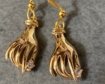 May include: Gold tone dangle earrings with a hand design and a small clear rhinestone in the center of each hand.