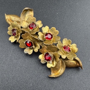 May include: Gold tone brooch featuring red rhinestone flowers. The brooch is shaped like a cluster of flowers with leaves.