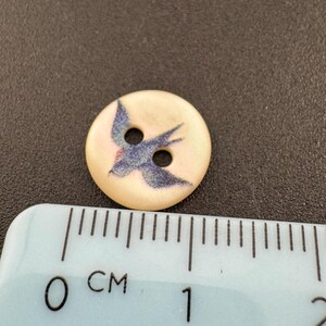 Pu&ograve; includere: Un bottone rotondo bianco sporco con un disegno di uccello blu. Il bottone ha due fori per cucire e misura circa 2 cm di diametro. L'uccello &egrave; raffigurato in volo, con le ali spiegate. Il bottone &egrave; probabilmente in plastica o un materiale simile.
