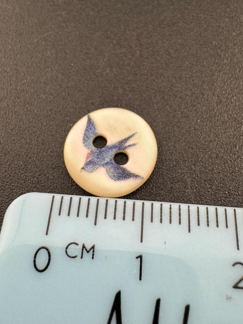 Pu&ograve; includere: Un bottone rotondo bianco sporco con un disegno di uccello blu. Il bottone ha due fori per cucire e misura circa 2 cm di diametro. L'uccello &egrave; raffigurato in volo, con le ali spiegate. Il bottone &egrave; probabilmente in plastica o un materiale simile.