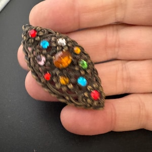 May include: An ornate, leaf-shaped brooch with a bronze-colored filigree design. The brooch is adorned with colorful, round gemstones in red, blue, yellow, green, pink, and white. The brooch is held in a hand.