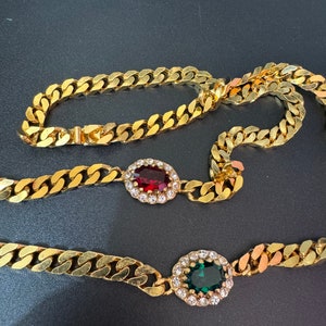 May include: A gold chain necklace with two oval-shaped pendants. One pendant features a red gemstone surrounded by clear rhinestones, and the other pendant features a green gemstone surrounded by clear rhinestones.
