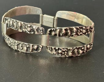May include: A silver-toned bracelet with a textured, organic design. The bracelet features a series of interconnected, rectangular links with a rough, uneven surface. The clasp is a simple, secure design. The bracelet is displayed against a dark background.