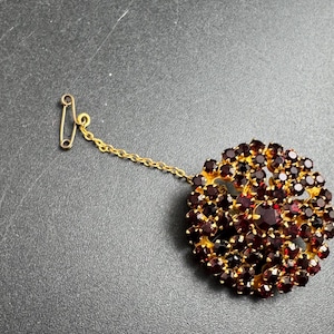 May include: A vintage brooch featuring a cluster of deep red gemstones set in a gold-toned metal frame. The brooch has a round shape with a safety pin clasp and a delicate chain. The gemstones are closely packed together, creating a rich, textured appearance.
