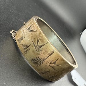 May include: A silver-coloured hinged bangle bracelet with etched bird and leaf designs. The bracelet has a rectangular shape and a small chain attached to the clasp. The etched designs are visible on the outer surface of the bracelet.