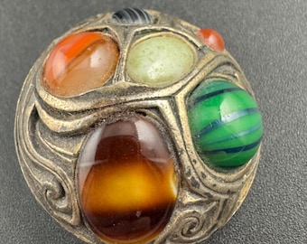 Può includere: Spilla vintage rotonda con cornice in metallo color oro antico. Presenta pietre cabochon arancioni, verdi, marroni e blu. Il design è ornato, con motivi a vortice. Un gioiello unico ed elegante.