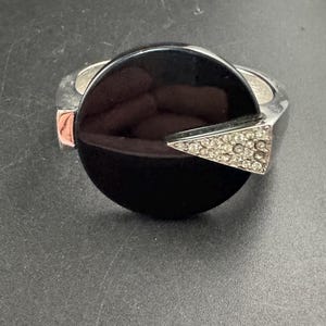 Könnte beinhalten: Ein silberfarbenes Gelenkarmband mit einem großen, runden, schwarzen Mittelstein. Ein dreieckiger Akzent, besetzt mit kleinen, klaren Kristallen, ziert den schwarzen Stein. Das Armband hat ein modernes, geometrisches Design.