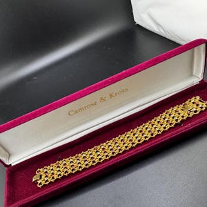 May include: A gold-tone bracelet with a delicate, openwork lattice design featuring small, embedded red and clear stones. The bracelet is presented inside a rectangular, hinged jewellery box lined with deep red velvet, marked Camrose & Kross.