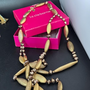 May include: A gold-coloured necklace featuring a variety of bead shapes, including textured ovals and small, faceted beads. The necklace is displayed with a pink Liz Claiborne box. The necklace has a lobster claw clasp. The necklace is a vintage piece.