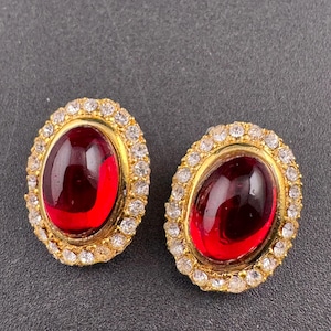 May include: A pair of oval-shaped earrings. Each earring features a large, deep red stone set in a gold-toned frame. The frame is surrounded by a halo of small, clear crystals. The earrings are displayed on a dark gray surface.