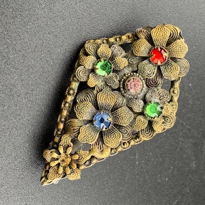 May include: A gold-toned brooch with a floral design. The brooch features five flowers with colourful rhinestones in the centre of each flower. The rhinestones are green, blue, red, and pink.
