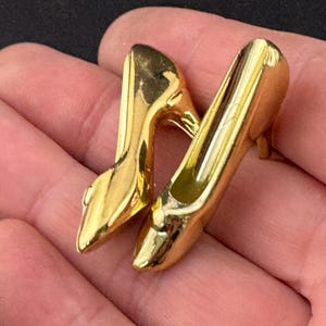 May include: A pair of gold-tone metal high heel shoe brooches. The shoes are in a classic style with a pointed toe and a high heel.