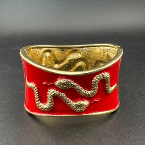May include: A wide hinged cuff bracelet featuring a vibrant red enamel finish. The exterior is decorated with a raised, textured gold-tone snake motif that wraps across the front. The interior of the bracelet has a polished gold-tone finish.