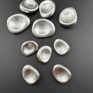 May include: A collection of ten silver-colored, pebble-shaped objects with a concave center. The objects are arranged on a dark surface. The smooth, metallic finish reflects light, creating highlights and shadows.