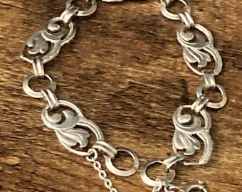 May include: A silver-toned metal bracelet featuring a series of repeating ornate, curved leaf-like links connected by small circular rings. The bracelet includes a delicate dangling chain segment and a circular spring ring clasp closure.