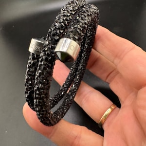 May include: A black wrap bracelet featuring a textured surface covered in small, dark, faceted crystals. The bracelet has two silver-toned metal end caps, with one cap clearly displaying the engraved text SWAROVSKI.