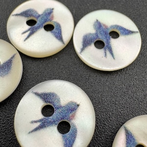 May include: A collection of round, off-white buttons. Each button features a blue and purple bird design with outstretched wings. The buttons have two holes for sewing and a glossy finish. Ideal for sewing projects.