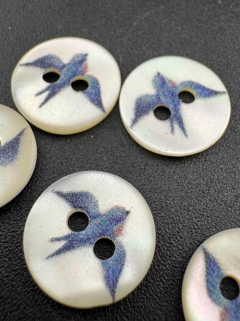 Pu&ograve; includere: Una collezione di bottoni rotondi, bianco sporco. Ogni bottone presenta un disegno di uccello blu e viola con le ali spiegate. I bottoni hanno due fori per cucire e una finitura lucida. Ideale per progetti di cucito.