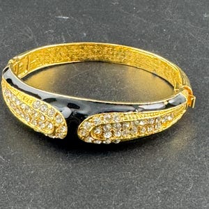 May include: Gold-toned hinged bangle bracelet with black enamel accents and clear crystal embellishments. The bracelet features a curved black enamel section with crystal accents on each side. The clasp is gold-toned.