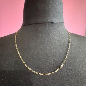 May include: A delicate gold necklace with a chain-link design. The necklace features small, round embellishments spaced along the chain. The jewelry is displayed on a black mannequin head against a pink background.