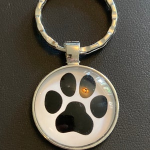 May include: A silver key chain with a round, white, glass charm featuring a black paw print.