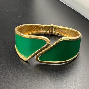 May include: A gold-toned hinged bracelet with a vibrant green enamel inlay. The bracelet features a unique, open design with a curved shape. The green enamel is framed by the gold metal, creating a striking contrast. The clasp is visible at the top.