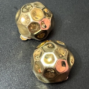 May include: A pair of gold-toned clip-on earrings with a geometric, faceted design. The earrings are round and have a textured surface.