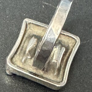 May include: Silver pendant with a square-shaped design featuring a textured surface and a raised design. The pendant has a loop at the top for attaching a chain.