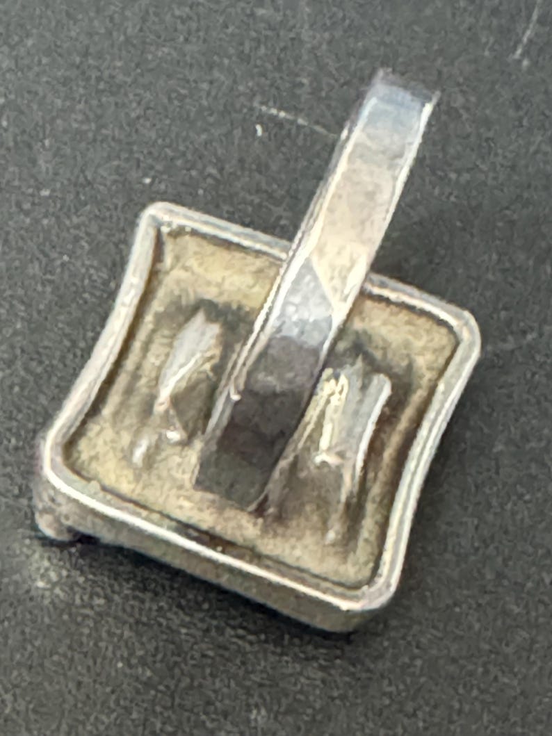May include: Silver pendant with a square-shaped design featuring a textured surface and a raised design. The pendant has a loop at the top for attaching a chain.