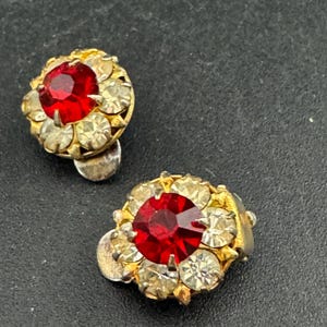 May include: A pair of vintage clip-on earrings. Each earring has a large, round, faceted red stone surrounded by smaller, clear, round stones. The setting is gold-toned metal. The earrings are on a dark background.