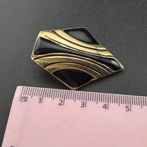 May include: A vintage brooch with a geometric design. The brooch features a black enamel inlay with gold-tone metal accents. The brooch is approximately 1.5 inches long.