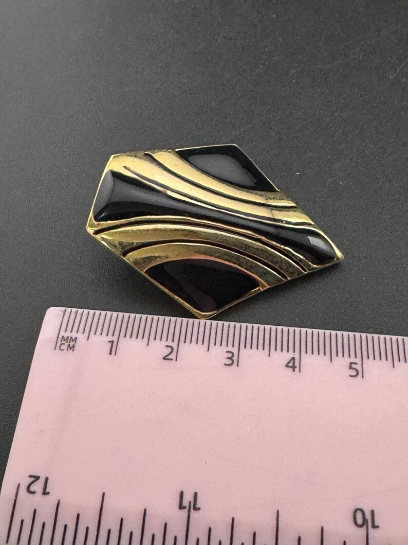 May include: A vintage brooch with a geometric design. The brooch features a black enamel inlay with gold-tone metal accents. The brooch is approximately 1.5 inches long.