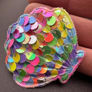 May include: A rainbow iridescent seashell-shaped patch with sequins. The patch is made of clear plastic with a white backing.