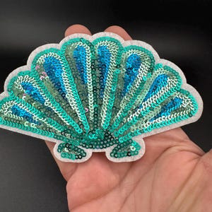 May include: A sequined seashell patch in shades of teal and blue, with a white felt backing. The sequins are arranged in a fan design. The patch is held in a hand. This decorative item could be used for embellishment on clothing or accessories.