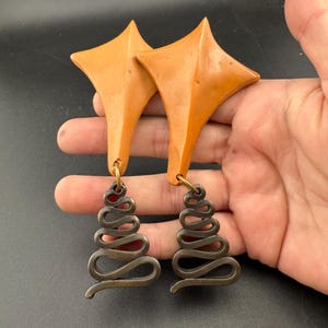 May include: A pair of distinctive earrings. Each earring has an orange star-shaped top and a bronze-coloured, spiral-shaped dangle. The earrings are suspended from a small gold-coloured loop. The background is a solid black.