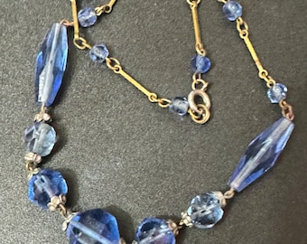 May include: A vintage necklace featuring a gold chain and blue glass beads. The beads are faceted and come in a variety of shapes, including round, oval, and rectangular.