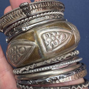 May include: A stack of multiple metal bangles and bracelets featuring various textures and designs. The collection includes wide cuffs with embossed floral patterns, twisted wire bands, smooth polished rings, and thin bands with serrated edges.