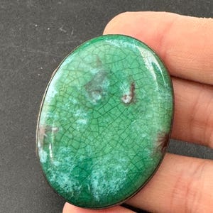 May include: Oval-shaped green and brown enamel pendant with a crackle effect. The pendant has a silver-colored metal border. The enamel has a glossy finish and is held in a hand.