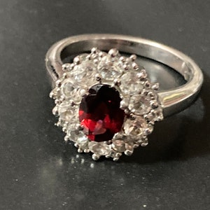 May include: A silver ring featuring a large oval red gemstone surrounded by smaller clear gemstones.