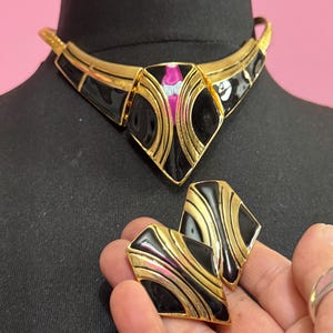 May include: A vintage gold-tone necklace and matching earrings set. The jewelry features black enamel and pink and white accents. The necklace is on a black mannequin head. The earrings are held in a hand.