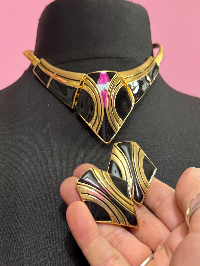 May include: A vintage gold-tone necklace and matching earrings set. The jewelry features black enamel and pink and white accents. The necklace is on a black mannequin head. The earrings are held in a hand.