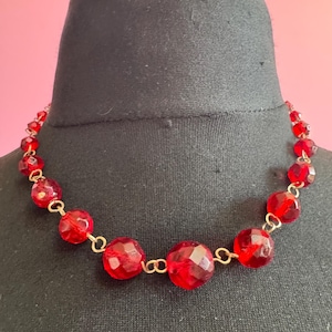 May include: A red beaded necklace with faceted beads and gold-coloured chain links. The necklace is displayed on a black mannequin head against a pink background. The beads are a deep, transparent red.