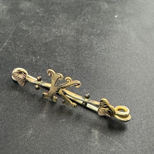 Peut inclure: Une épingle de barre dorée avec des accents floraux et de feuilles. L'épingle présente un élément décoratif central et de petits détails en relief le long de la barre. L'épingle est probablement vintage ou antique.