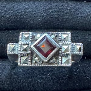 May include: A silver ring featuring a central square-cut deep red garnet stone set in a diamond orientation. The rectangular face of the ring is decorated with multiple small, faceted marcasite stones arranged in a geometric pattern around the center.