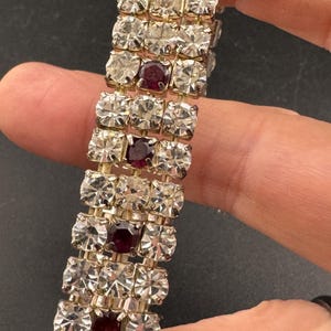 May include: A vintage bracelet featuring rows of clear, square-cut rhinestones alternating with rows of deep red, square-cut stones. The bracelet has a gold-tone metal setting and is shown against a dark background.